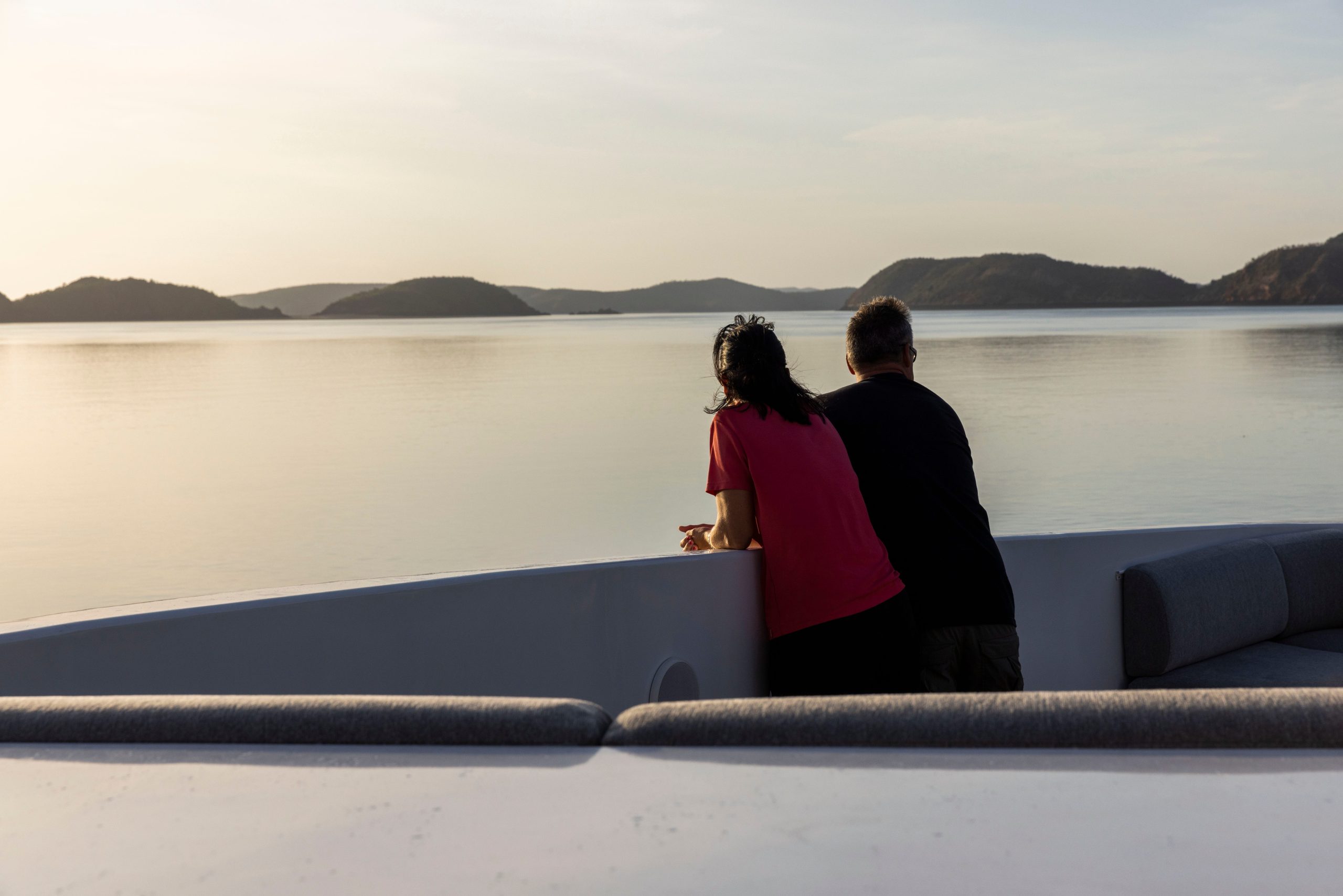 Two guests lean peacefully on the bow of the TRUE NORTH expedition vessel, gazing across calm golden waters as the sun sets over the Kimberley coastline. 