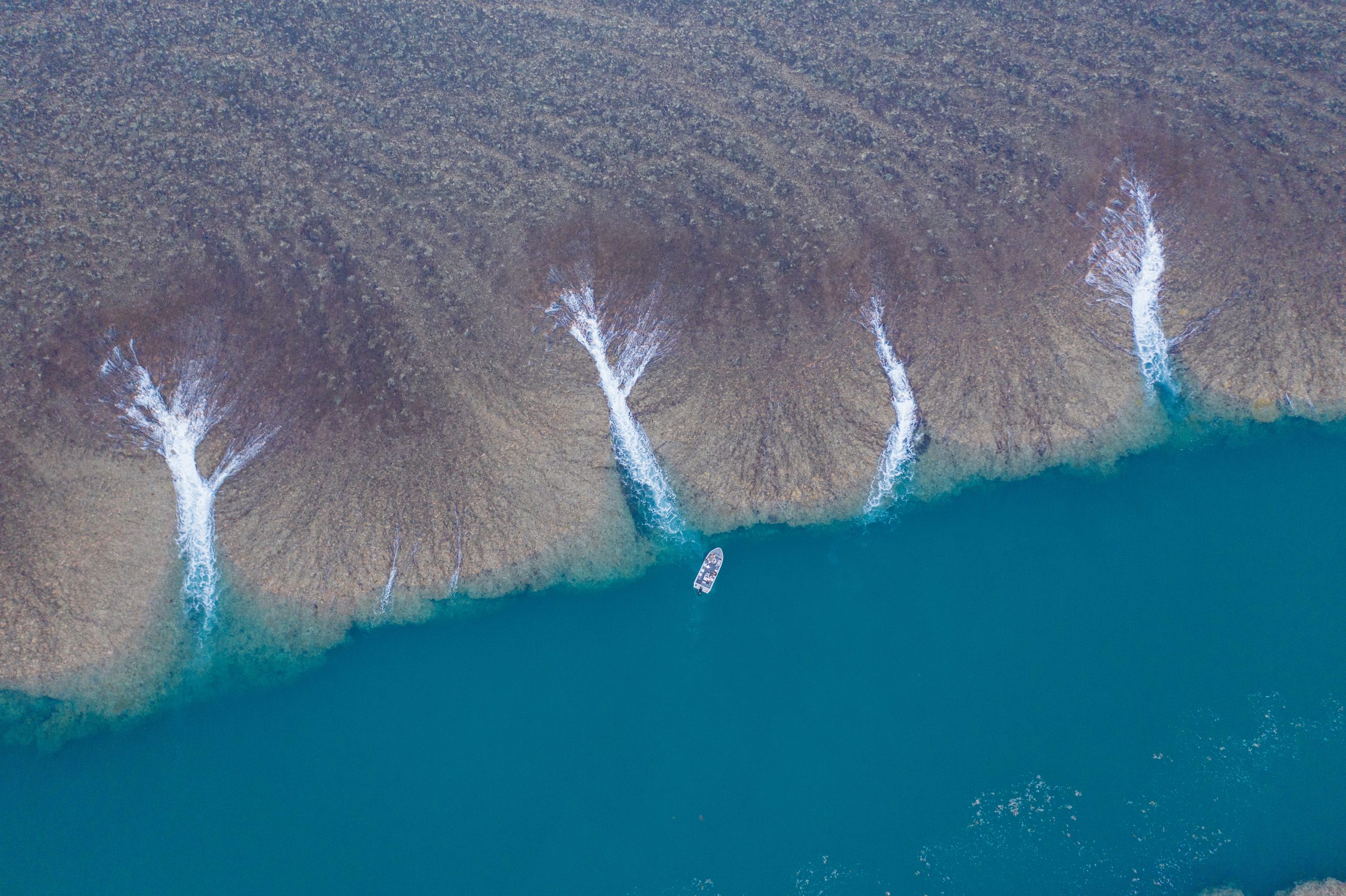 Aerial view of a True North adventure boat navigating along Montgomery Reef, where waves break in white patterns against shallow coral formations and meet calm, deep blue water, illustrating remote coastal exploration in pristine conditions.