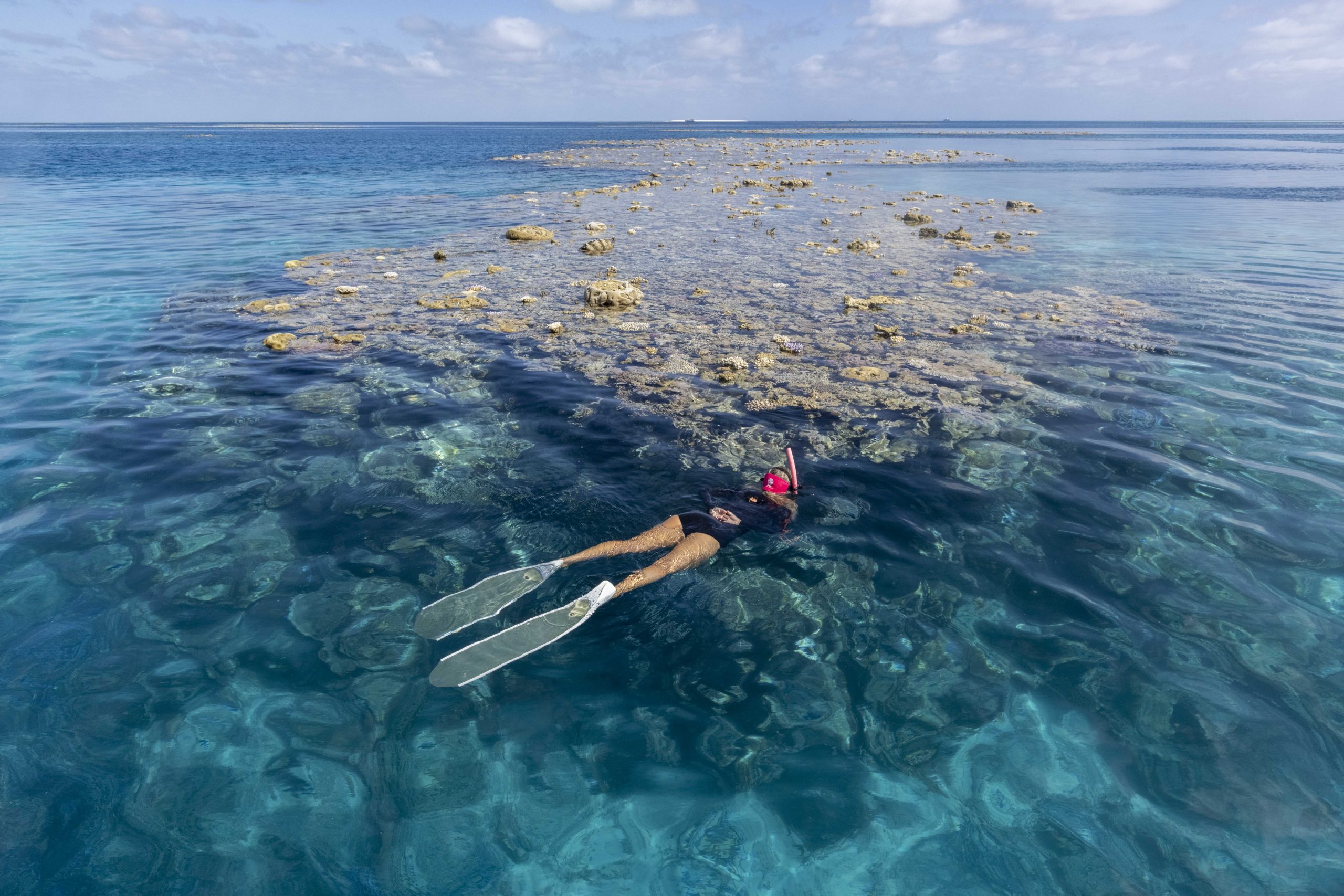 Snorkeller floats in crystal-clear waters near a shallow coral reef on the Great Barrier Reef, surrounded by vibrant blue ocean and scattered coral heads.