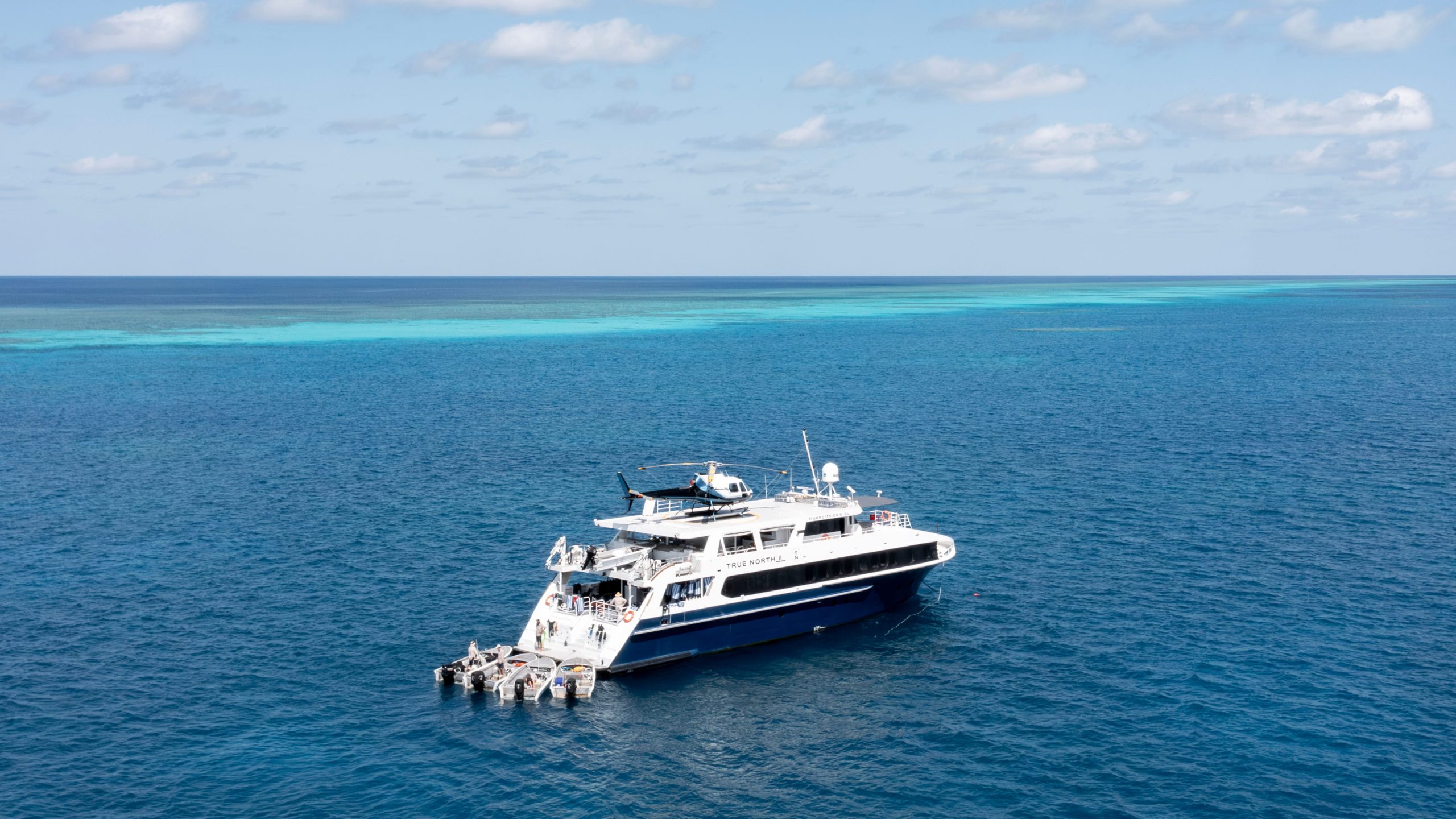 Luxury expedition vessel TRUE NORTH II anchored in the turquoise waters of the Great Barrier Reef, with a helicopter on its deck and tender boats moored at the stern.