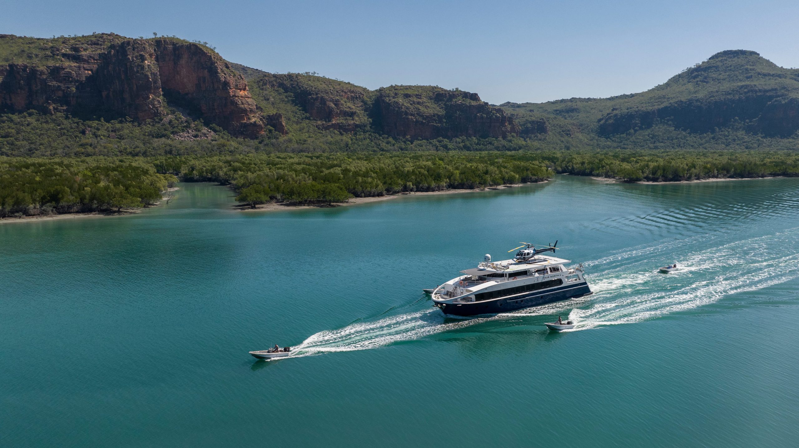 TRUE NORTH cruising through a Kimberley river system with adventure boats exploring nearby mangroves and cliffs.