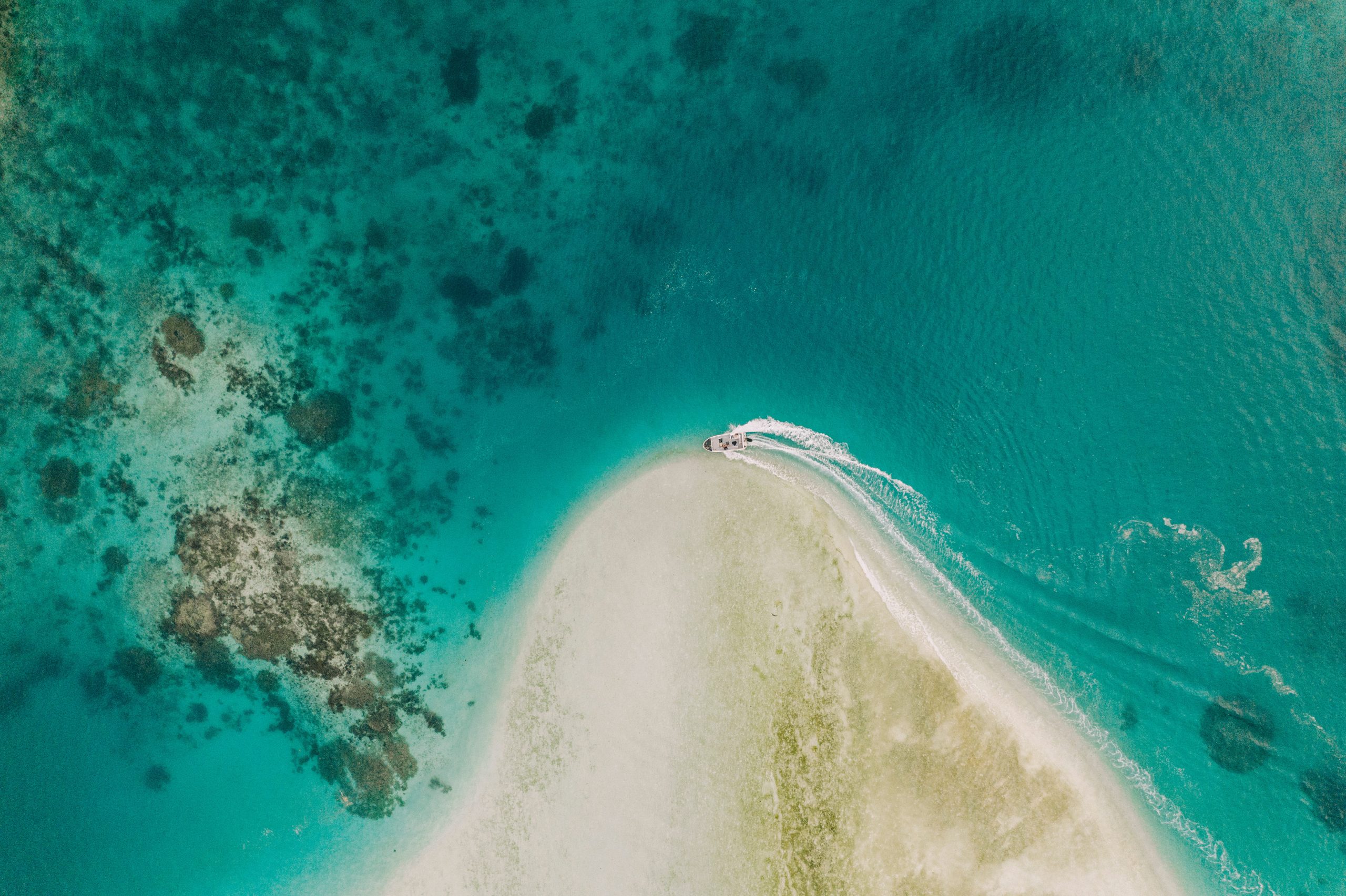 Aerial view of a small boat approaching a remote sandbar surrounded by vibrant turquoise waters and coral reefs in Raja Ampat, Indonesia