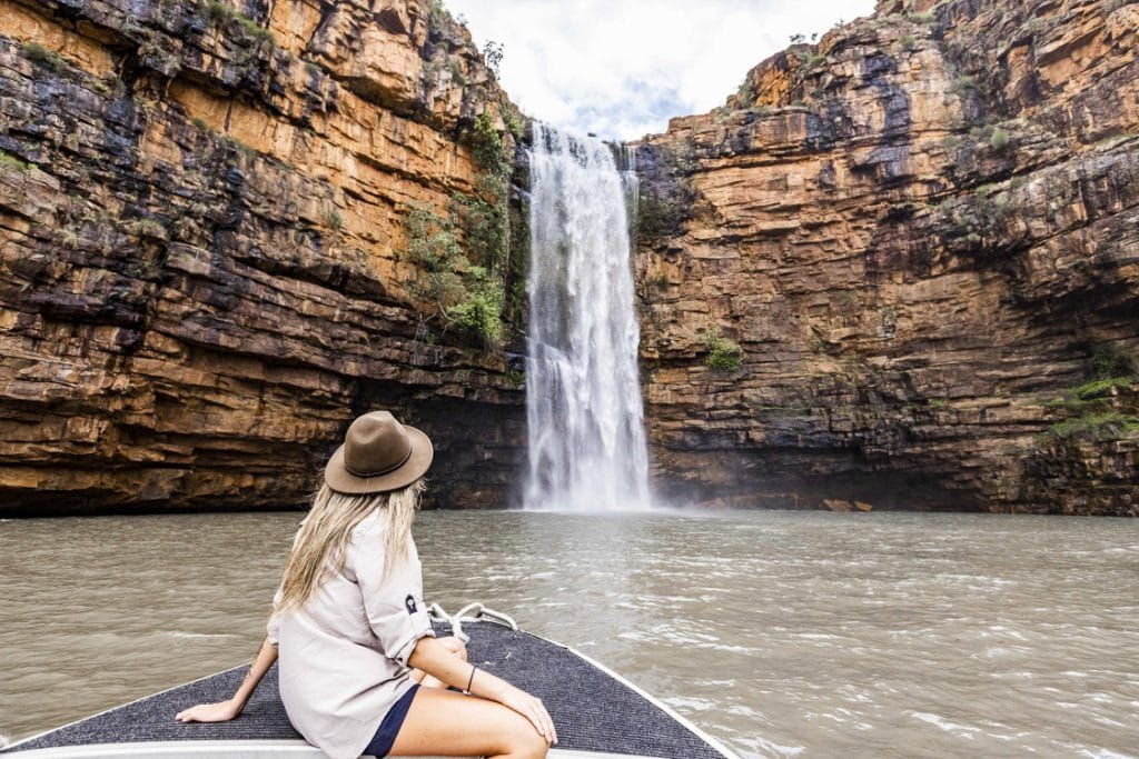 Woman sitting alone in a small boat, looking up at a towering cliff with water cascading over the edge