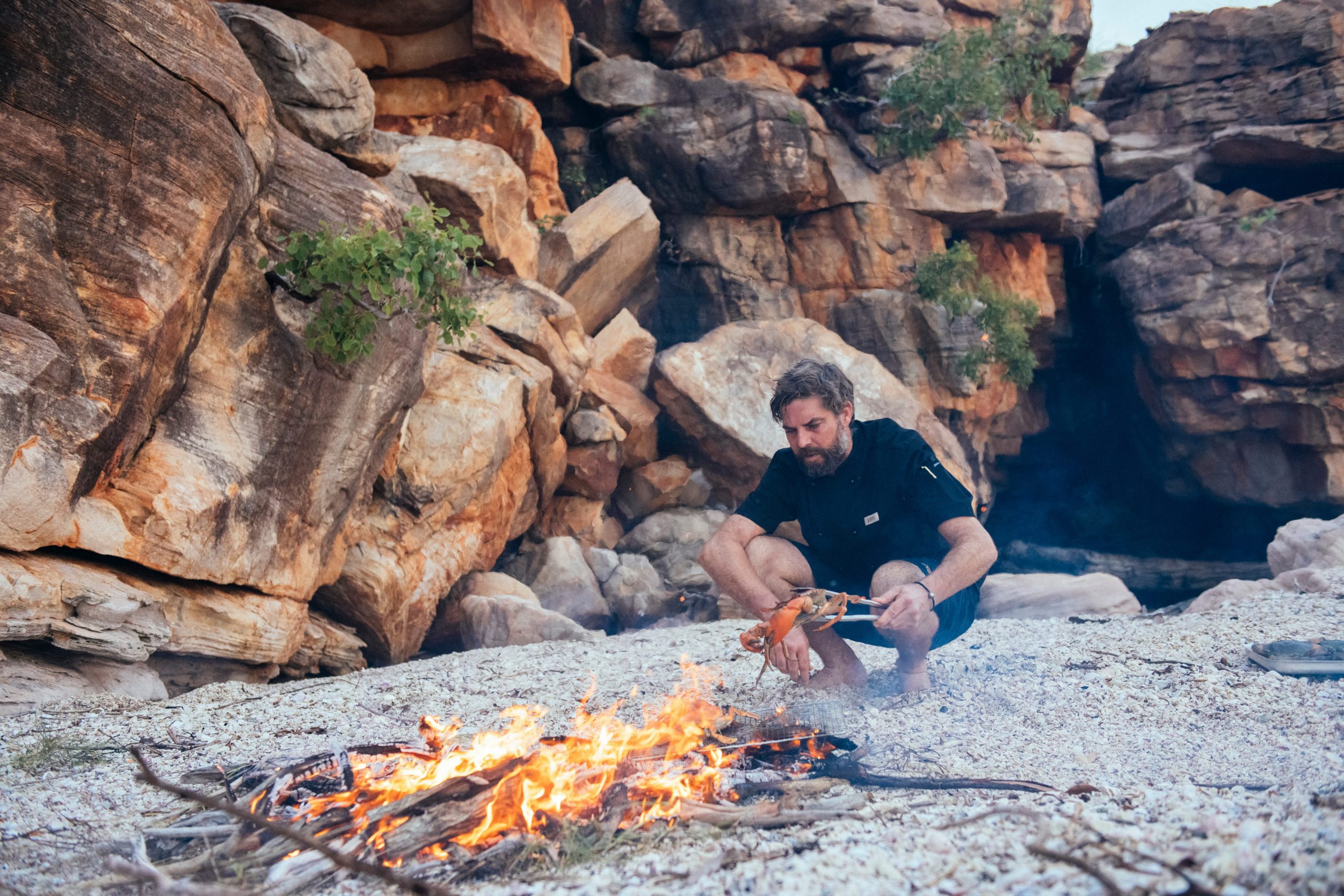 Crew member preparing freshly caught seafood over an open fire on a remote Kimberley beach, reflecting the immersive, expedition-style experiences of a True North journey.