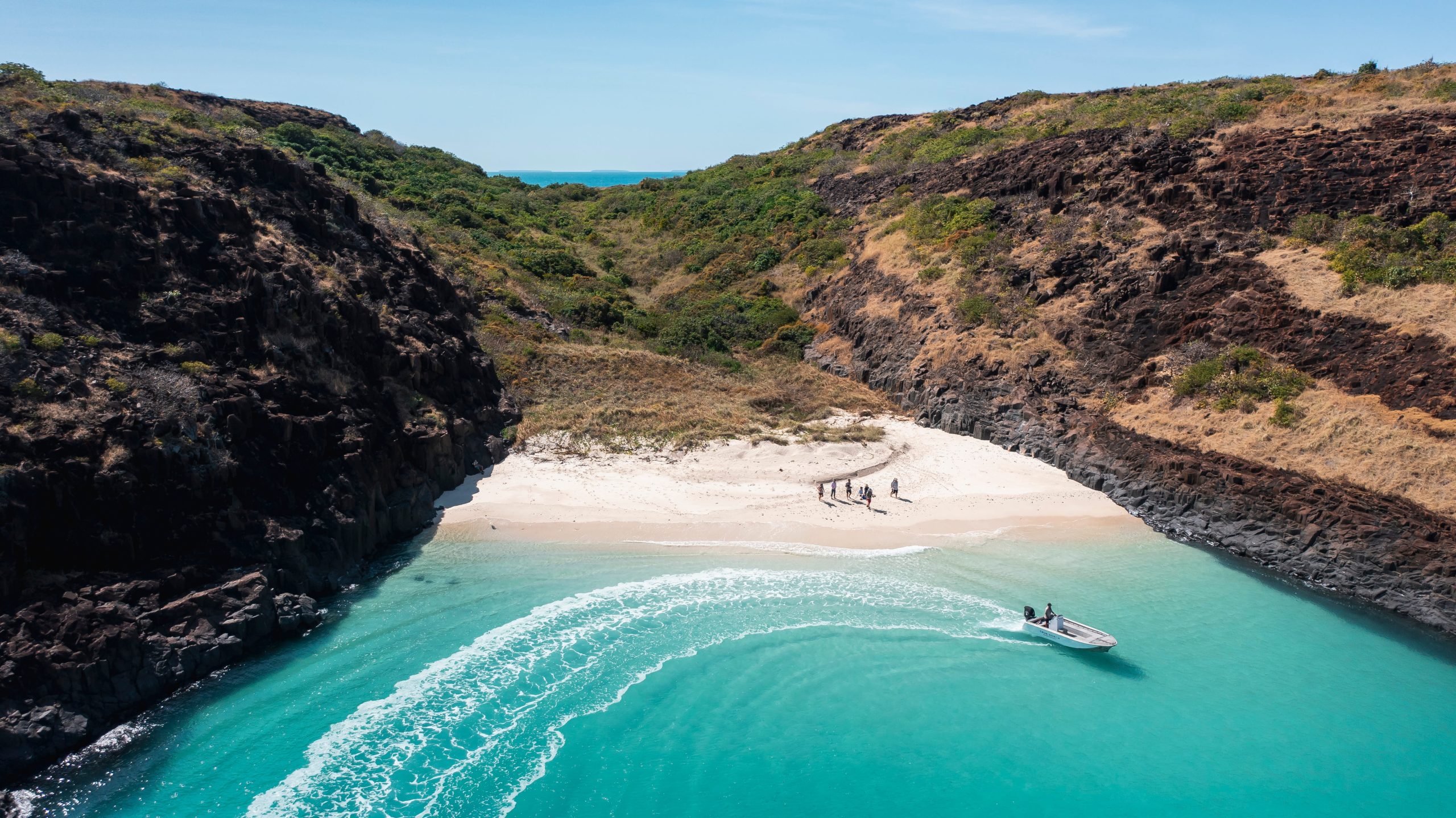 Aerial view of a secluded turquoise bay framed by rugged cliffs, with a tender approaching a pristine beach, highlighting the remote access and untouched beauty of a True North expedition. 