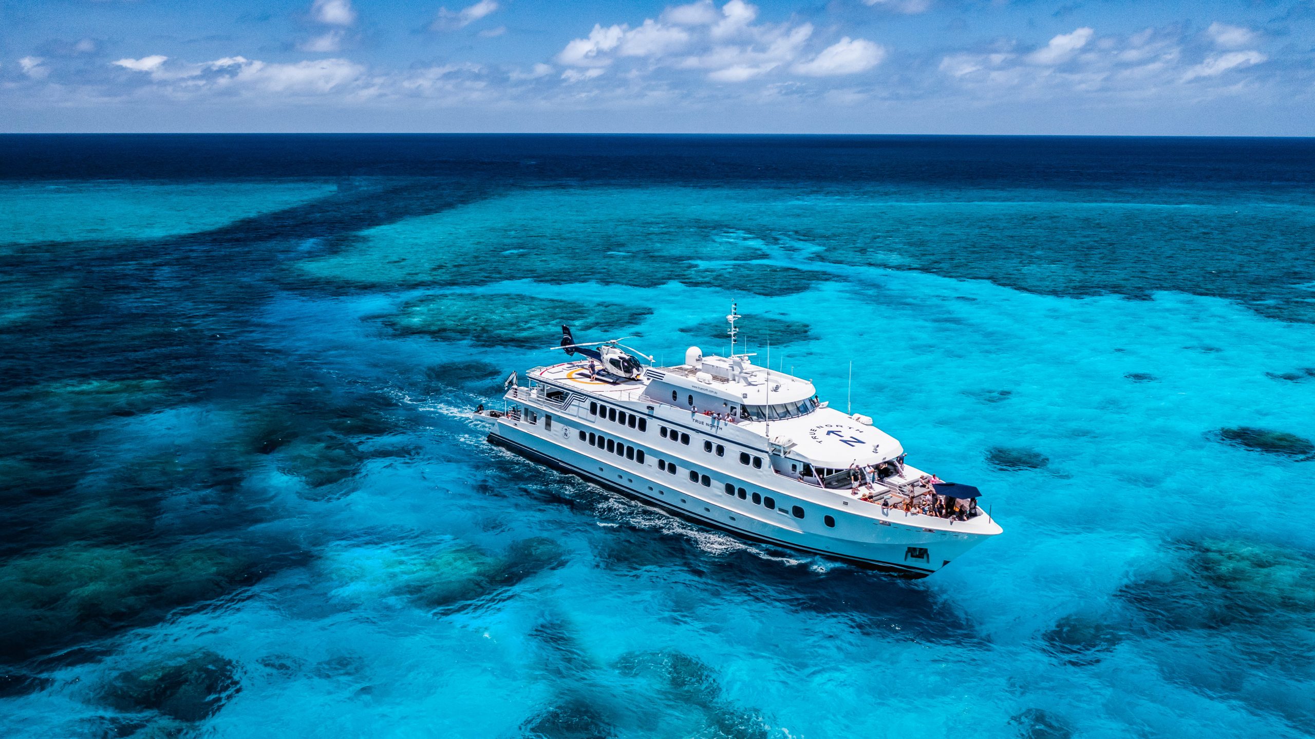 Aerial view of the True North sailing through clear turquoise waters over a coral reef, with a helicopter on deck, illustrating the calm conditions and remote coastal exploration of shoulder season travel in Australia.