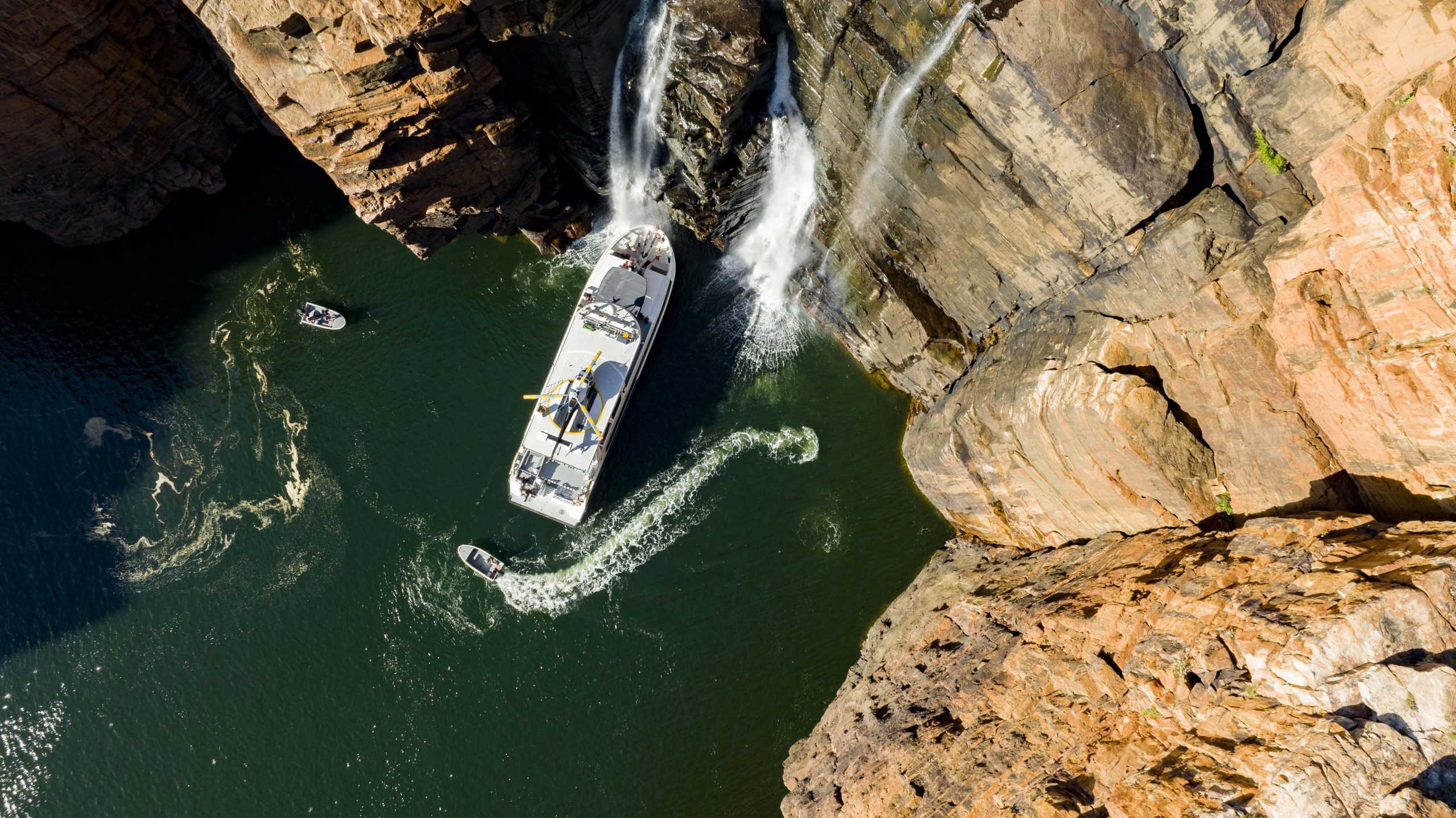 Aerial view of a small expedition cruise vessel navigating a narrow gorge beneath cascading waterfalls, highlighting the access and calm conditions of shoulder season travel in remote Australia.