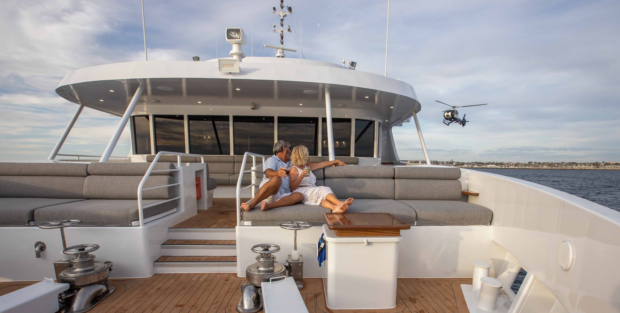 Couple relaxing on the deck of a True North luxury cruise with helicopter flying nearby.